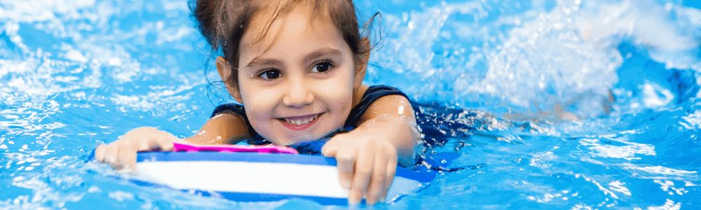 Zainteresowania małego dziecka - uśmiechnięta dziewczynka w wieku przedszkolnym pływa w basenie z pomocą deski