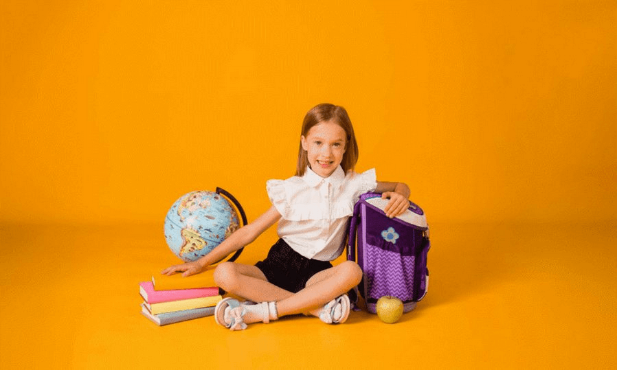 Plecak dla pierwszoklasisty - jaki wybrać? Uśmiechnięta dziewczynka z plecakiem i wyprawką szkolną siedząca na podłodze